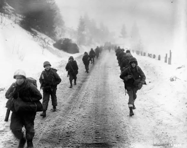 Photo of a column of 16th Regiment troops advancing on a snowy road during the Battle of the Bulge.